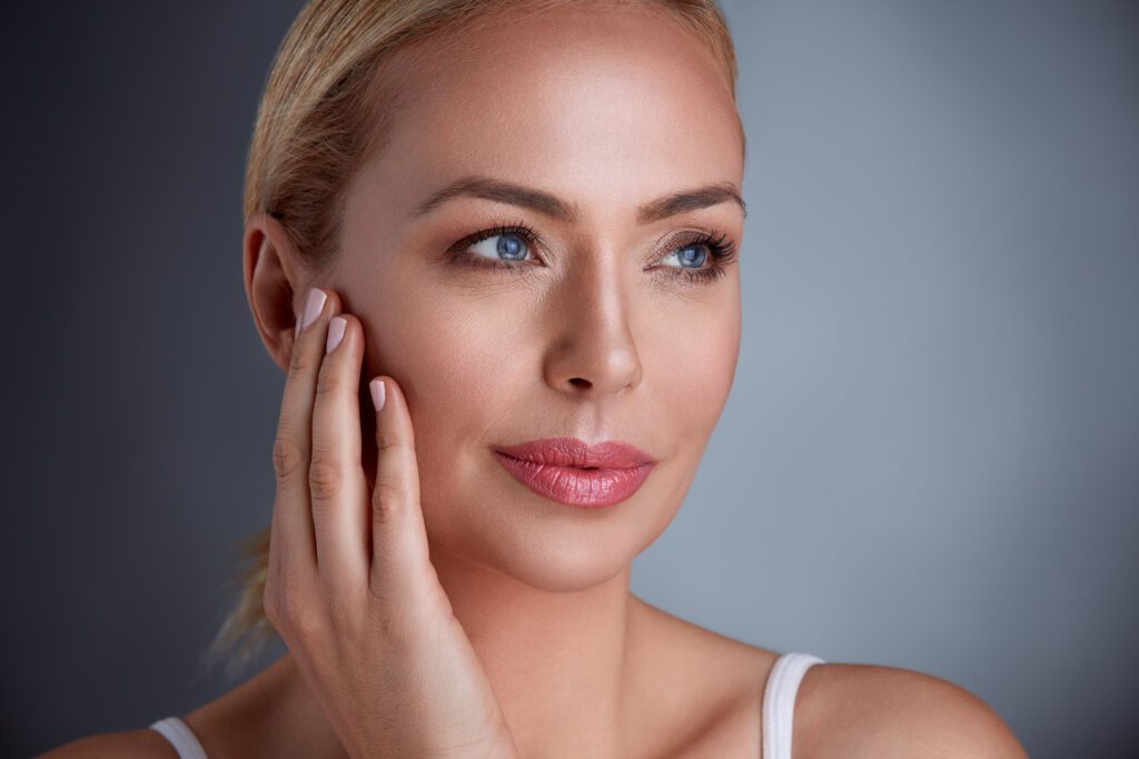 A woman with blonde hair and blue eyes touching her face.